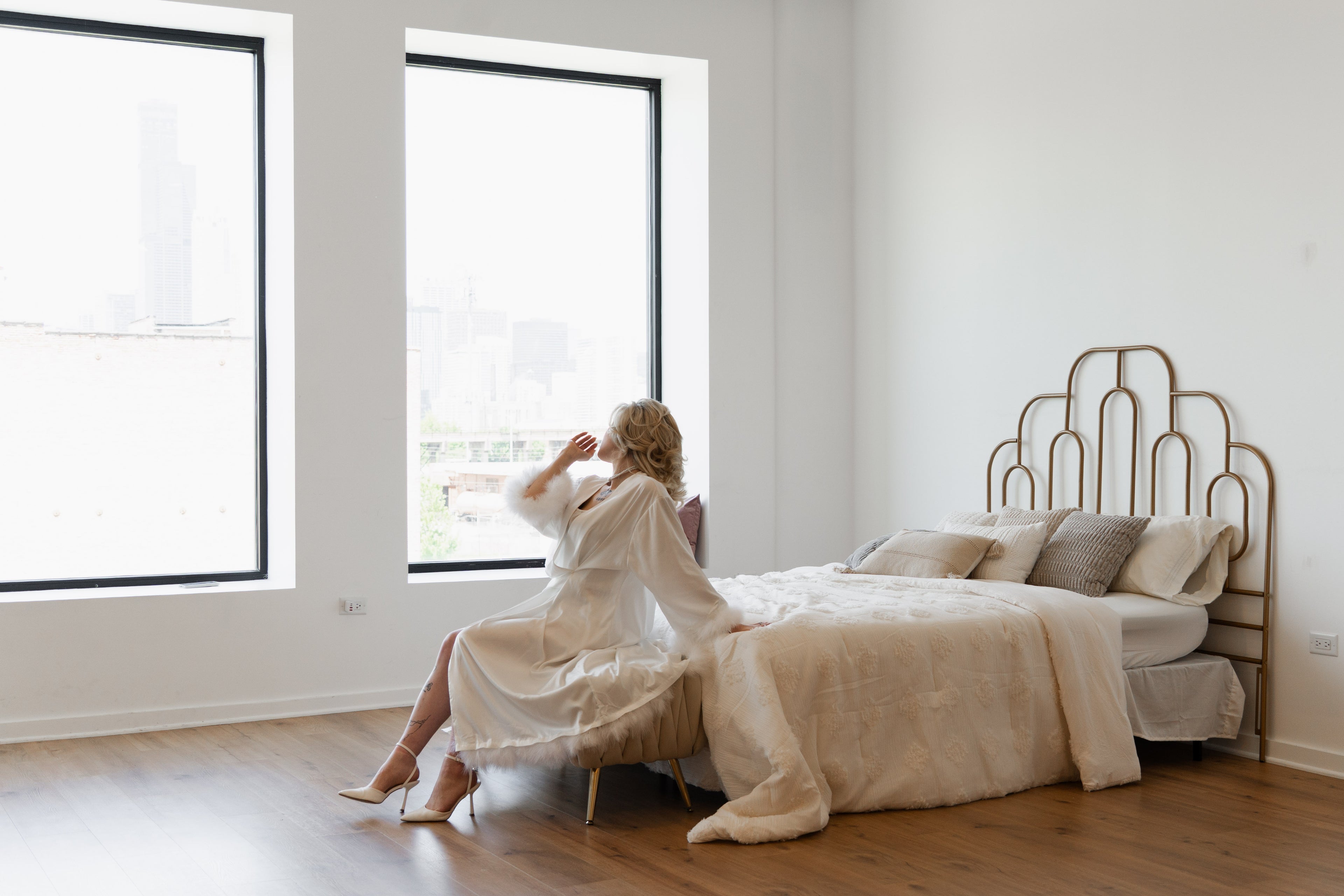 Woman in a robe sitting on the floor next to a bed in a bright bedroom.