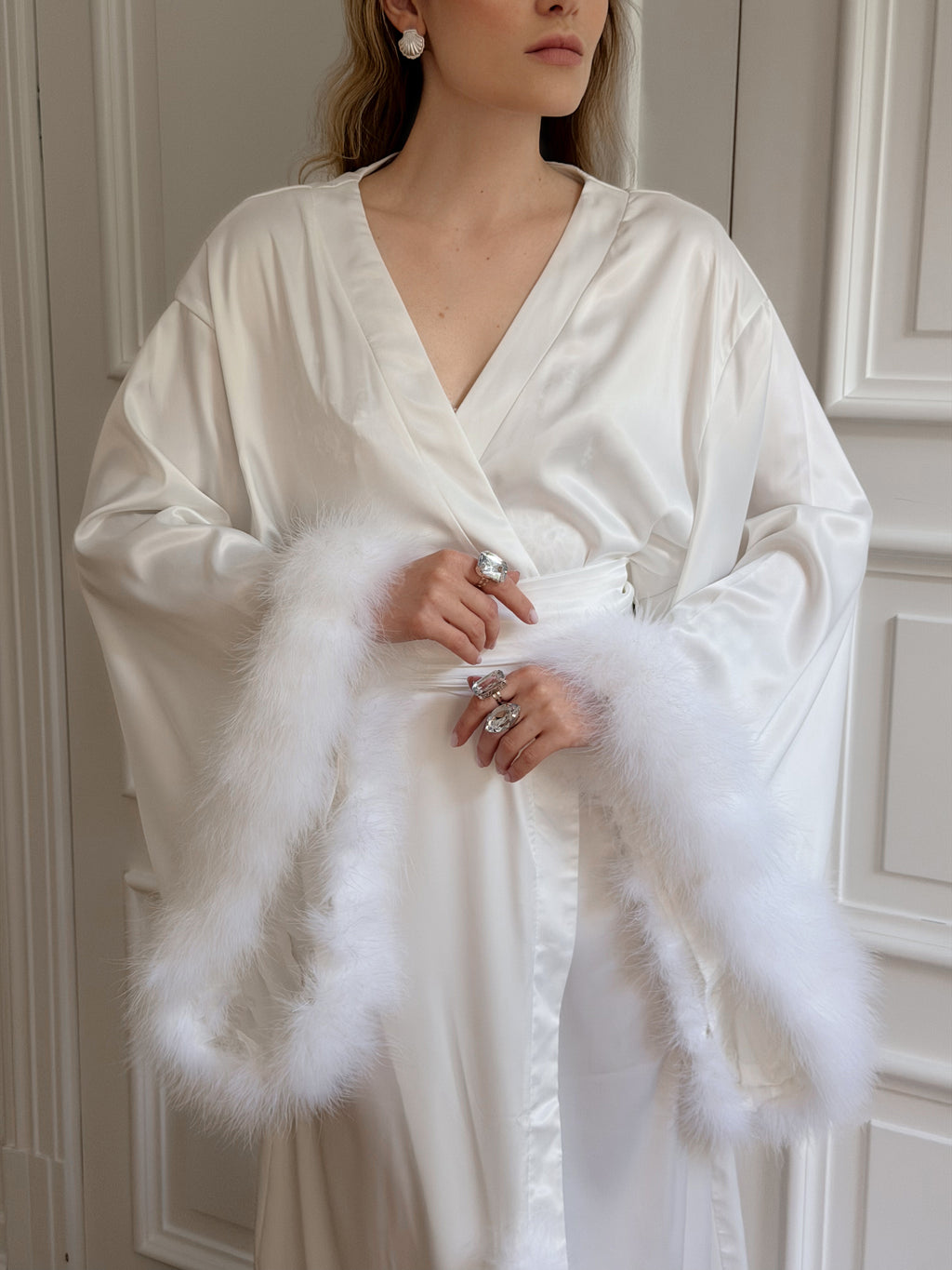 A woman wearing a white satin robe with fluffy white feather trim along the sleeves and waist, standing indoors against a light paneled wall.