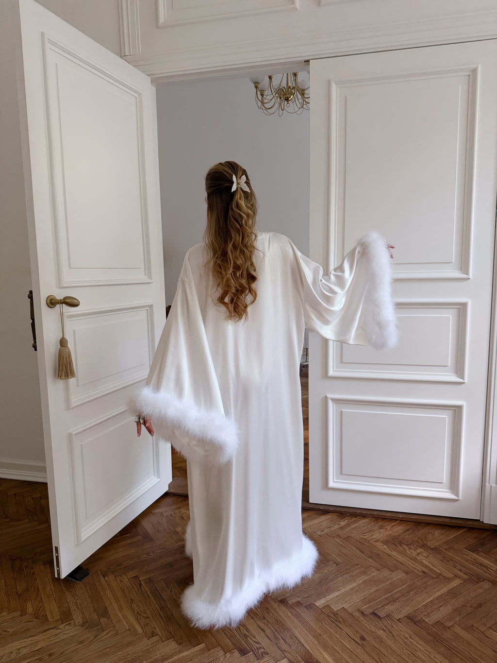 A woman viewed from behind wearing a long white robe with fluffy white trim at the hem and sleeves, standing in an open doorway on a wood floor.