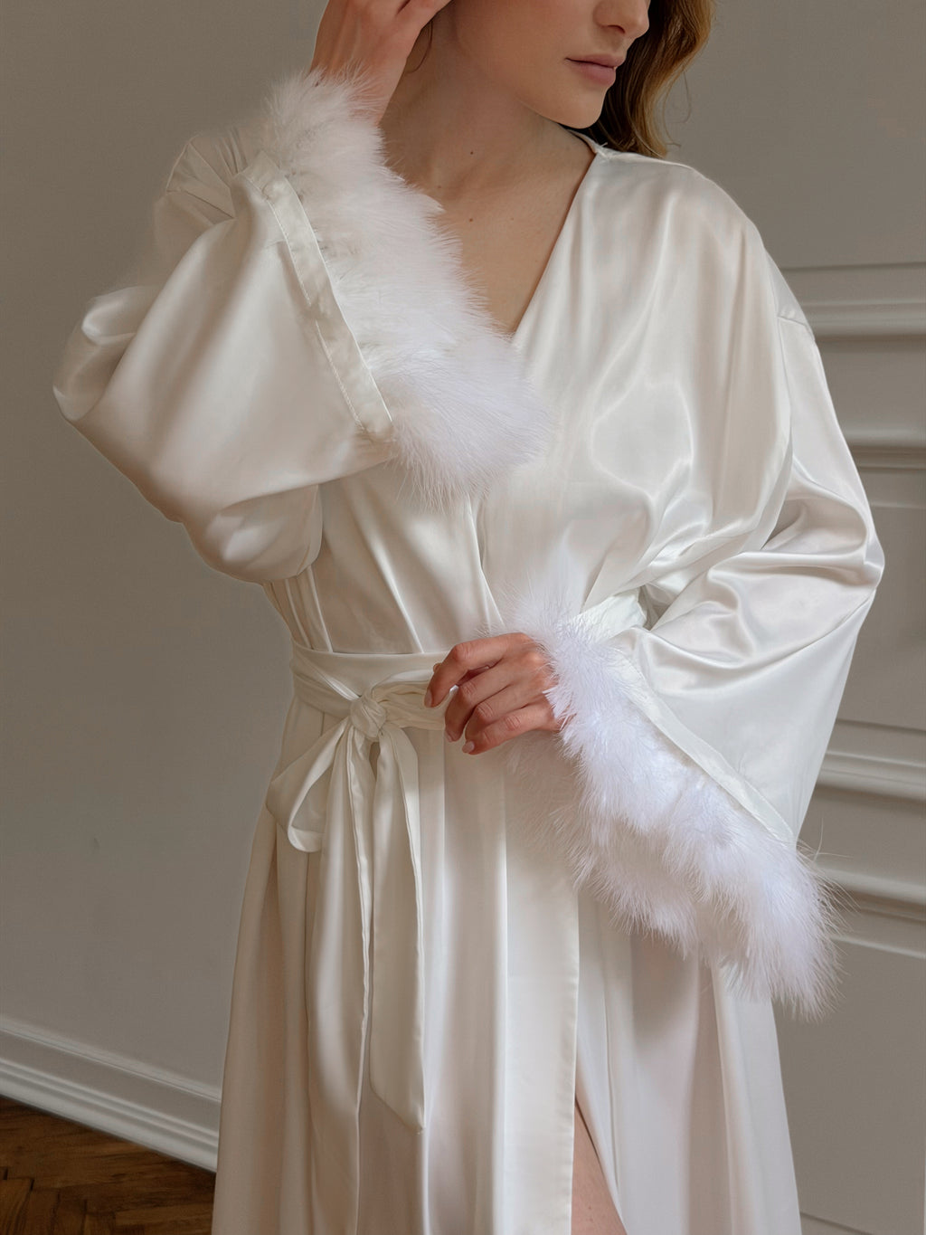 A woman wearing a satin robe in warm ivory or cream, featuring fluffy white faux-fur trim along the collar and cuffs, with a matching tie belt. The photo is taken in a softly lit indoor setting with a neutral wall background.