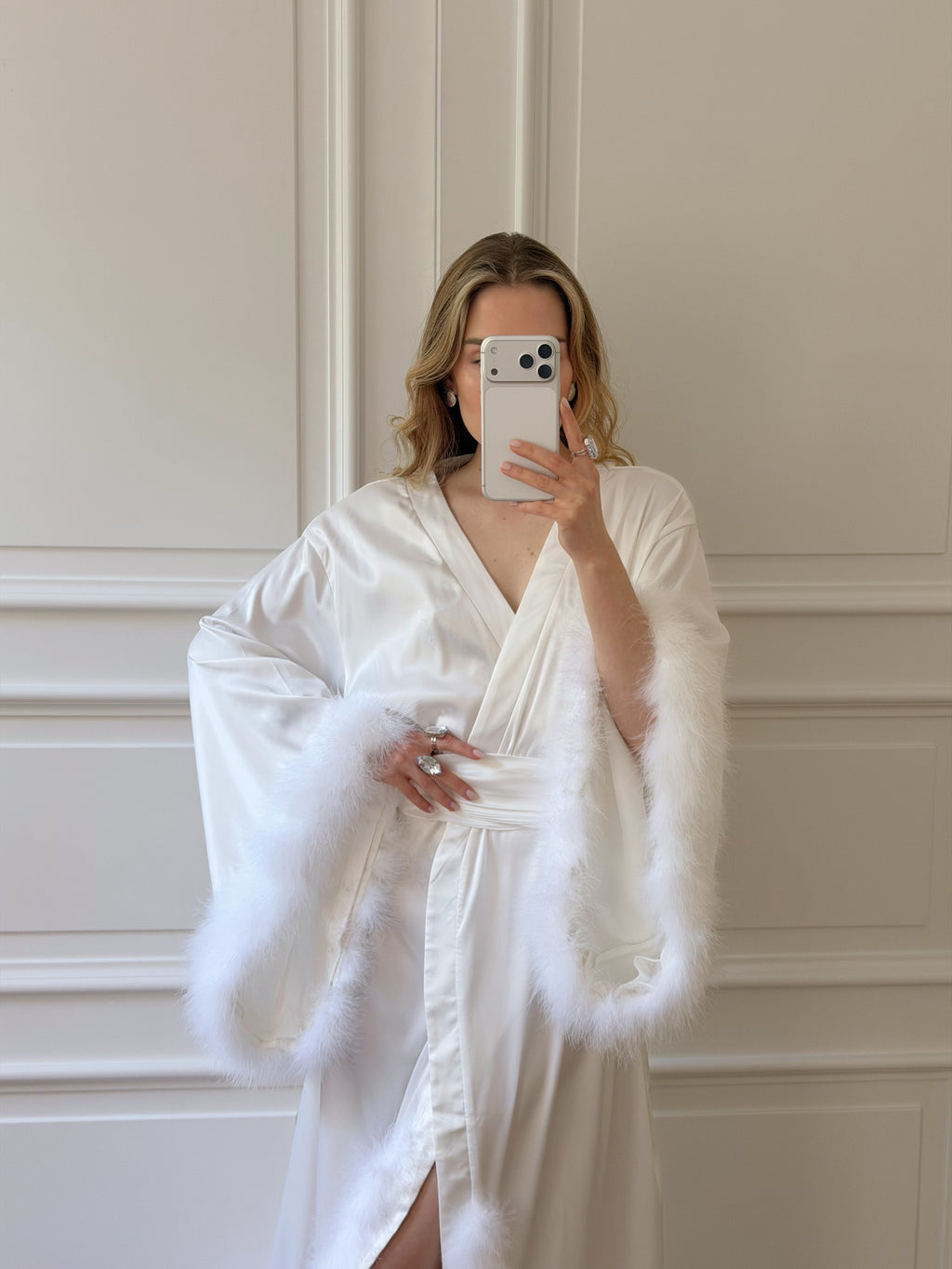 A woman takes a mirror selfie wearing a white satin robe with fluffy white faux-fur trim, standing against a white paneled wall.