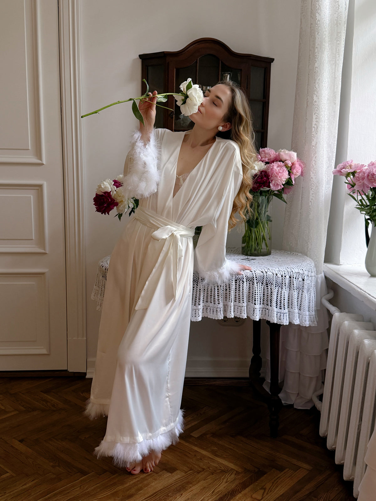 A woman wearing a long cream satin robe with white feather trim stands barefoot in a vintage room, holding a white rose near her face. A lace-covered table with pink flowers and a dark wooden cabinet are behind her.