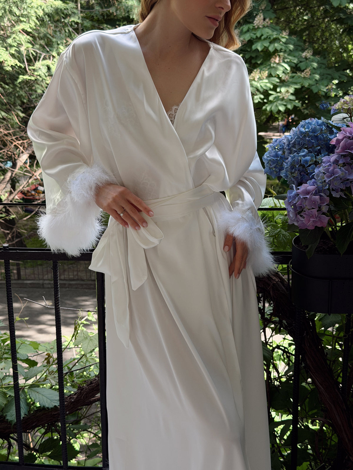 A woman wearing a white satin robe with fluffy faux-fur trimmed cuffs, standing outdoors on a balcony with greenery and flowers in the background.