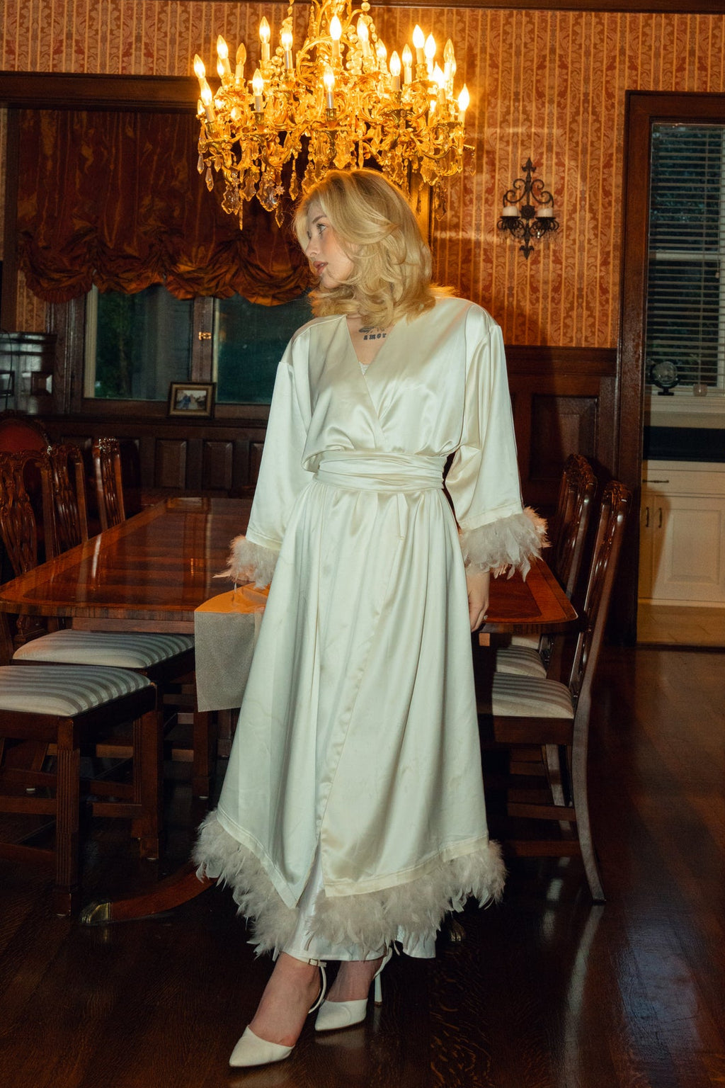 Woman in a white dress standing in a room with wooden furniture and a chandelier.