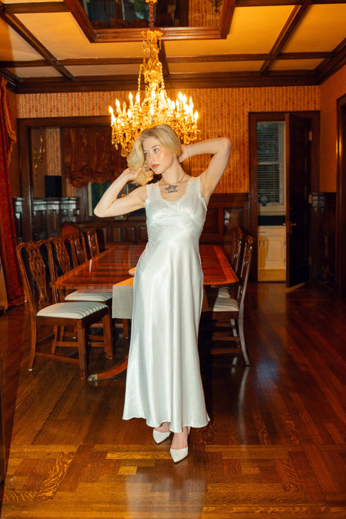 Woman in a white dress standing in a warmly lit dining room with wooden floors and a chandelier.