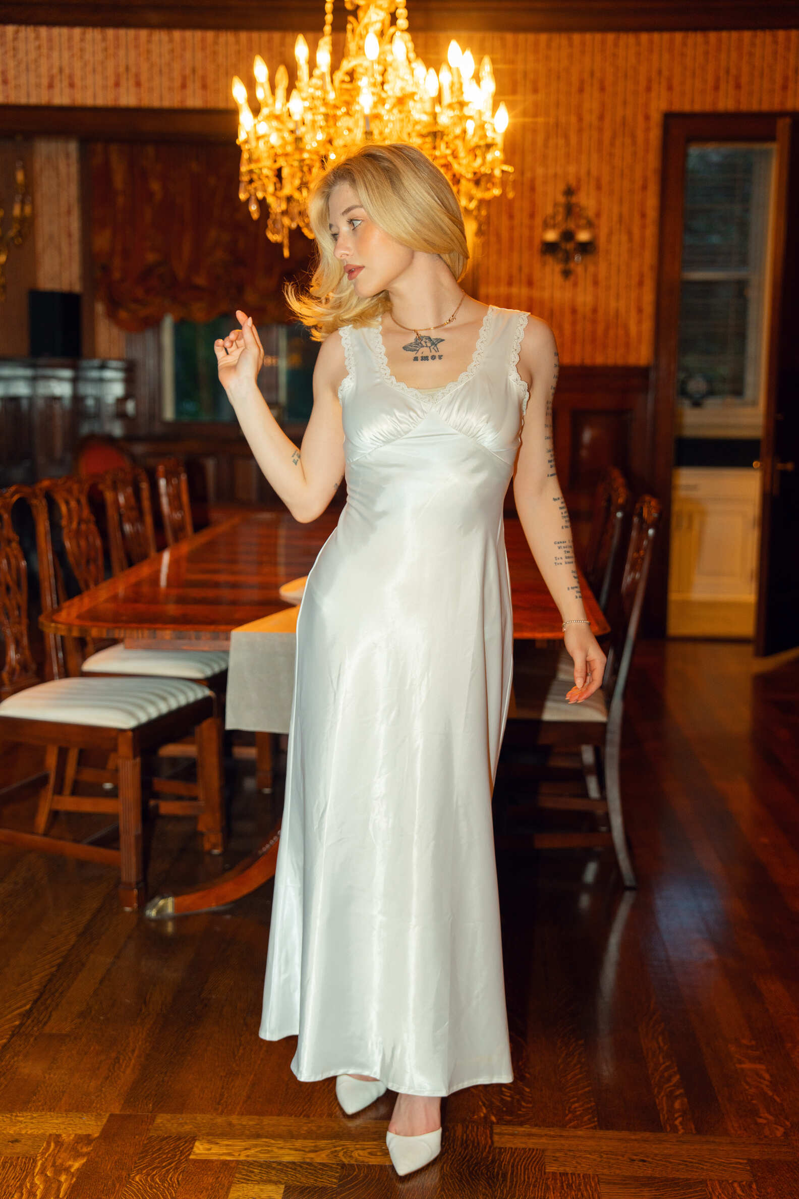 Woman in a white dress standing in a room with wooden furniture and a chandelier.