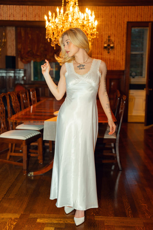 Woman in a white dress standing in a room with wooden furniture and a chandelier.