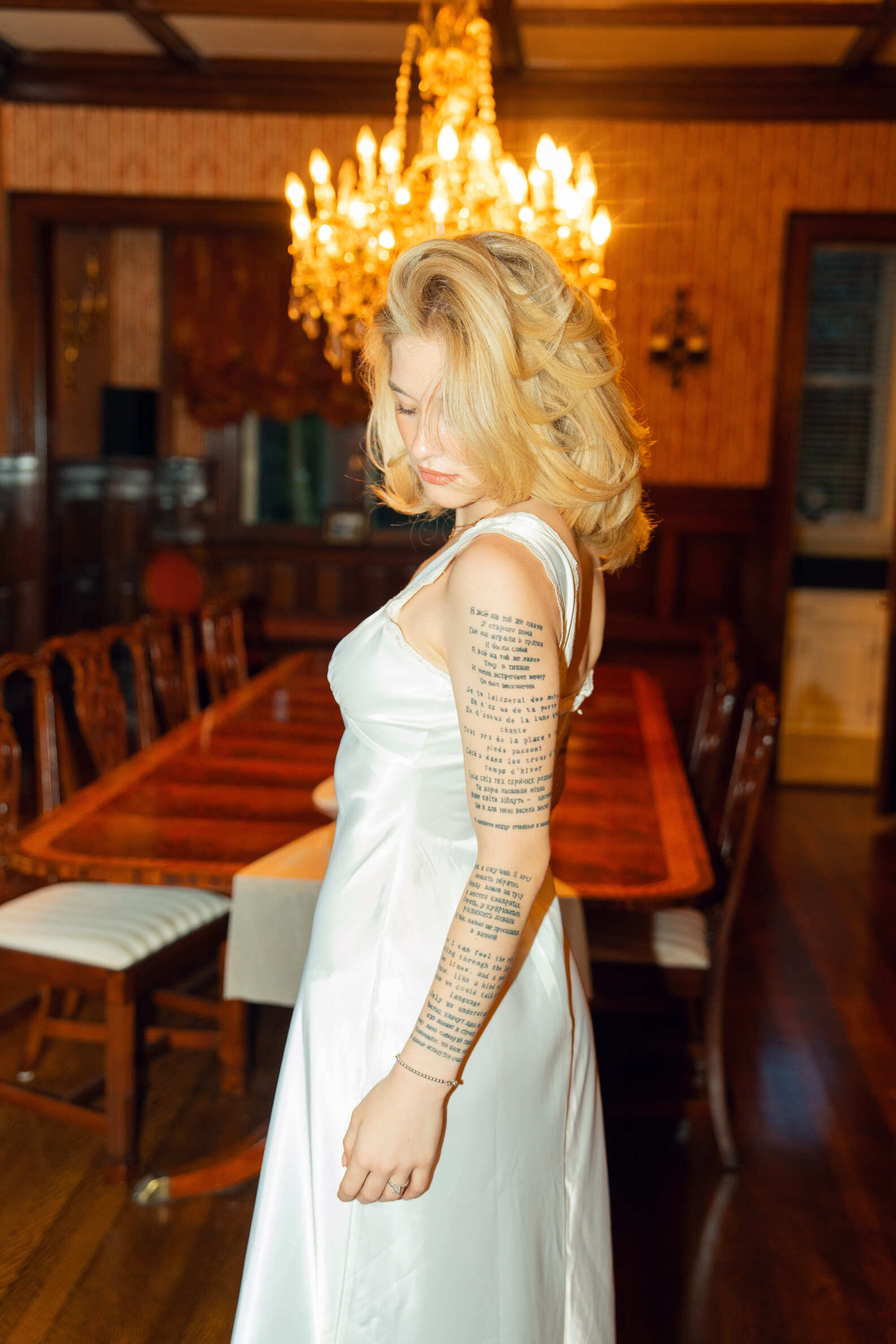 Woman in a white dress with tattoos standing in a room with wooden furniture and a chandelier.