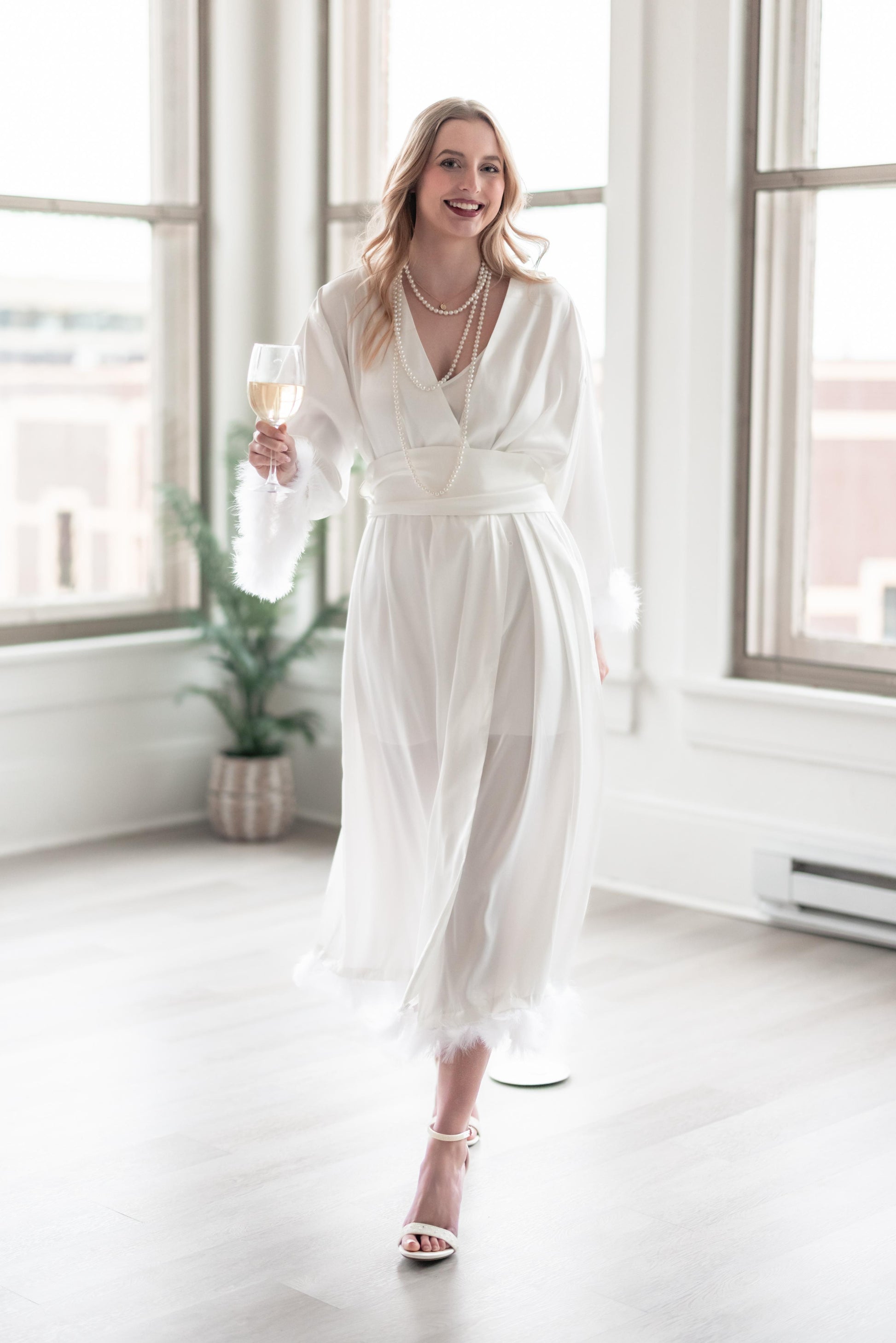 Woman in a white dress holding a glass of champagne in a bright room with large windows.