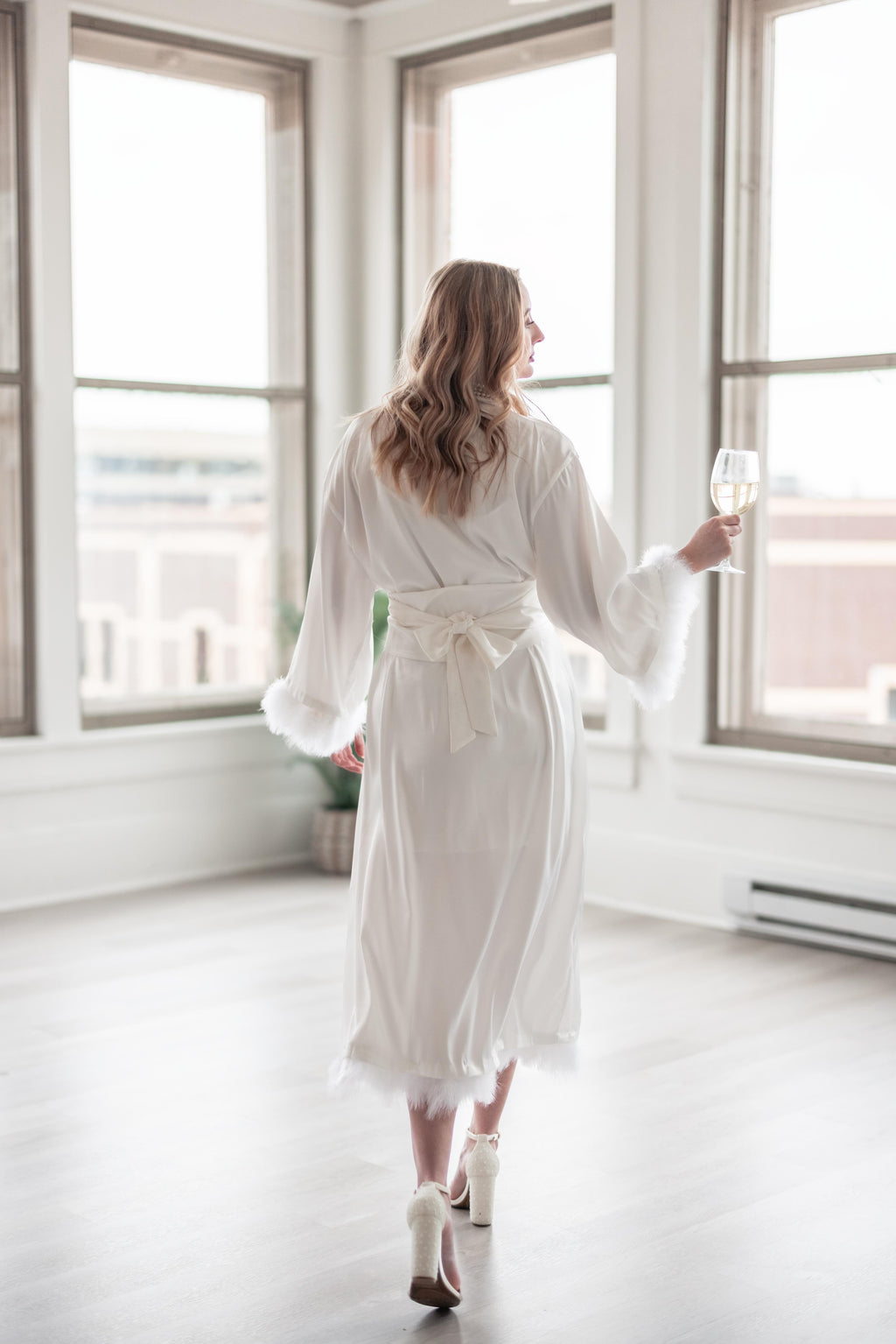 Woman in a white robe holding a glass of wine in a bright room with large windows.